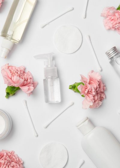 Top view of different cosmetic bottles, carnations flowers, cotton sticks and cosmetic pads on white