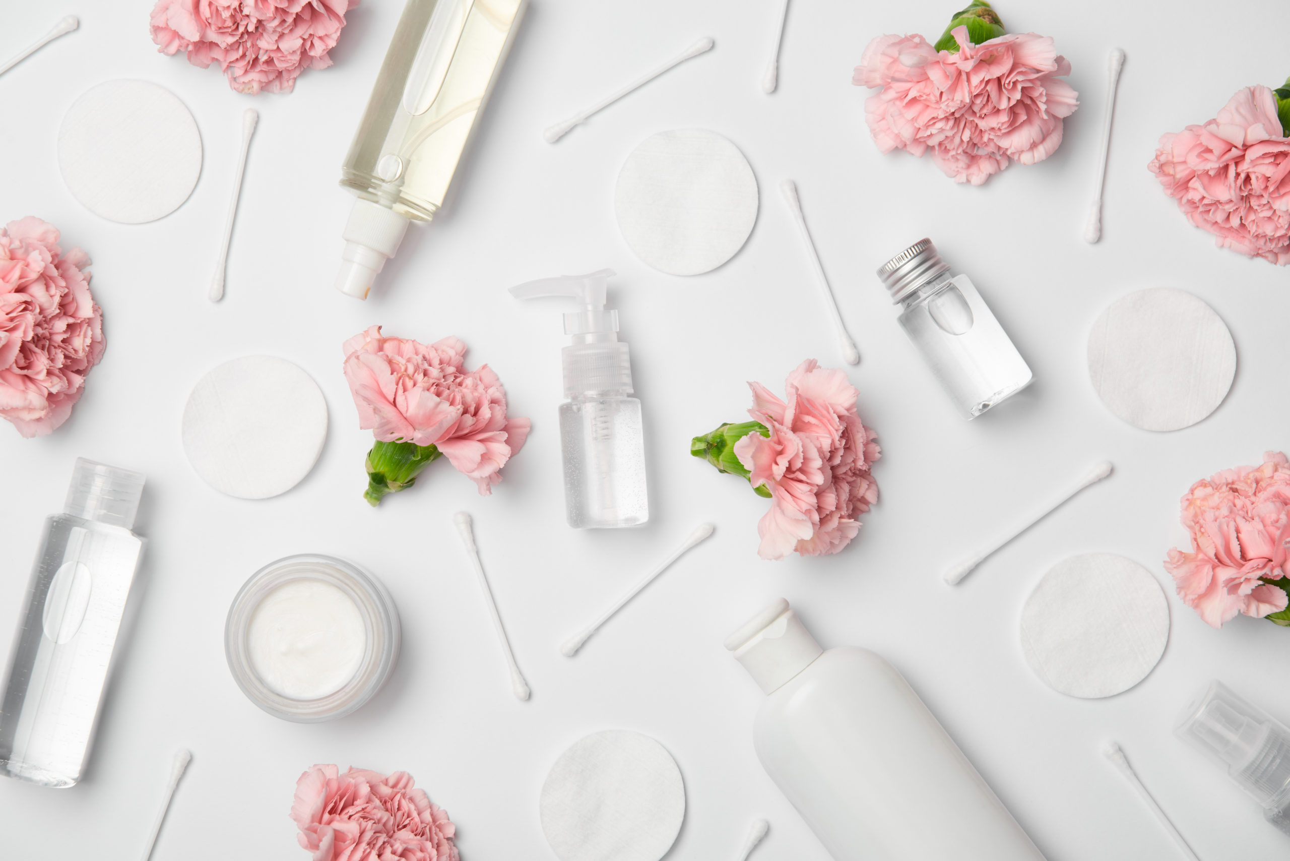 Top view of different cosmetic bottles, carnations flowers, cotton sticks and cosmetic pads on white